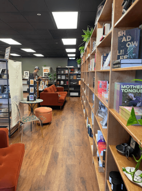 A cozy bookstore interior with wooden shelves filled with books, a reading area, and a person browsing in the background.